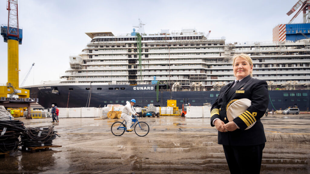 Captain Inger Klein Thorhauge standing in front of the Queen Anne cruise ship at a shipyard.